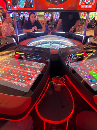 This image depicts the interior of a casino on the Hollywood Seminole Reservation in the United States. The primary focus is an electronic roulette table, featuring a central roulette wheel surrounded by individual player consoles. Each console has a touchscreen display showing betting options and game information. Red LED lighting illuminates the table's edges and features. A partially consumed drink sits on the table. In the background, other patrons are seen at various gaming machines. The lighting suggests a nighttime or indoor environment. The scene conveys a casual atmosphere with people engaged in gambling activities.