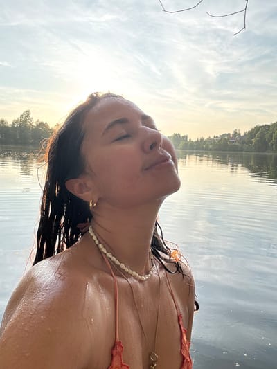 A young woman with wet, dark hair is shown from the chest up, leaning her head back with her eyes closed, facing the sky. She is in calm, rippling water, possibly a lake, with trees and buildings visible on the distant shoreline. The sun is low in the sky, casting a warm glow and creating a lens flare. She is wearing a beaded necklace and a coral-colored bikini top. Her skin is glistening with water droplets. There are no other people or obvious signs of city life in the immediate vicinity. The location is identified as the city of Kamysh, Russia, though this cannot be visually confirmed from the image.