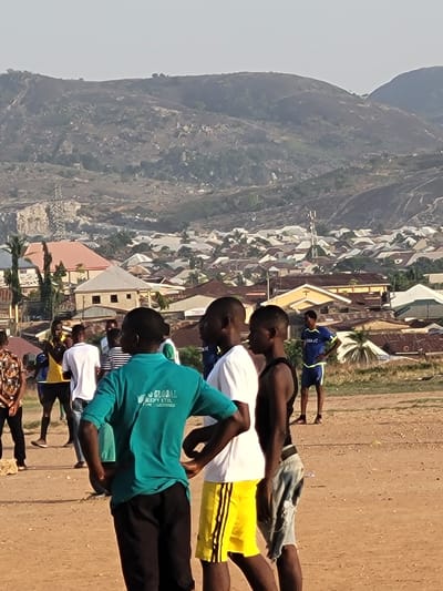 The image depicts a group of people standing on a dirt field in Ibo, Nigeria. In the background, there is a hillside and buildings. The people are wearing a variety of clothing, including t-shirts and shorts. Some of them appear to be exercising or stretching. The overall scene suggests an outdoor activity or gathering in a residential area near a natural landscape feature.