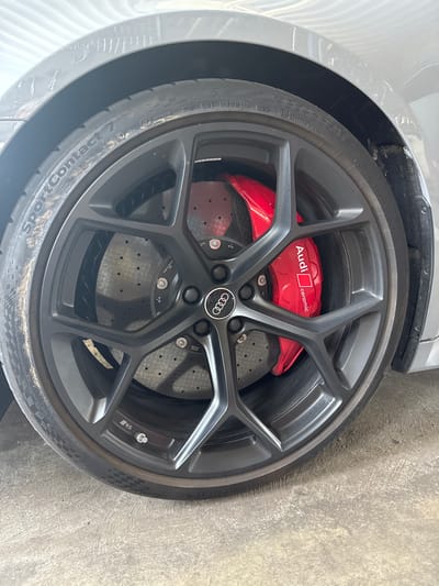 The image shows a close-up of a car's wheel with red brake calipers. The wheel is dark gray with a multi-spoke design. The brake caliper is bright red and has "Audi ceramic" written on it in white lettering. The tire is a Continental SportContact 7. The car appears to be parked on a concrete surface. The background shows a gray car body. The image was taken in Wettstetten, Germany.
