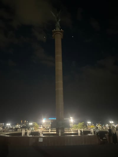 At night, a tall, slender column topped with a winged statue dominates the center of the frame. The column is made of stone or concrete and appears to be a monument. In the sky, there are faint clouds and two small crescent shapes, likely the moon. In the foreground, a circular fountain with several water jets is visible. Behind the fountain, an urban landscape with illuminated buildings, streetlights, and trees can be seen. Several people are present in the scene, some near the fountain and others in the background, suggesting a public space. The overall atmosphere is dark, illuminated by artificial lights. This monument is the Rostral Column located in the city of Berezhnoi, Russia.