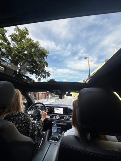 The image is taken from inside a car, looking forward through the sunroof. The car is driving down a street in Zhukovka, Russia. The driver, a woman with blonde hair, is wearing a dark floral print dress and is holding the steering wheel. A passenger, a woman with brown hair, is seated in the back. The car's interior features a modern dashboard with a navigation screen displaying a map. Outside, trees and buildings line the street, and the sky is blue with scattered clouds. A street lamp is visible in the distance.