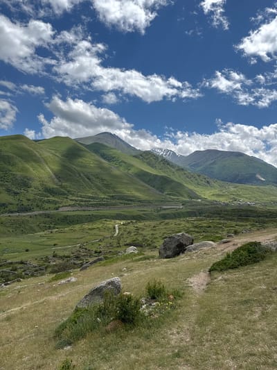 The image depicts a wide, scenic mountain landscape, presumably in El'tiubiu, Russia, under a bright blue sky dotted with fluffy white clouds. The foreground is characterized by a sloping, grassy hill with scattered large rocks and a visible dirt path leading into the distance. The midground features rolling green hills and valleys, with a road or railway line running horizontally across the scene. In the background, larger, more rugged mountains rise, some of which have patches of snow on their peaks. A small village or settlement is visible nestled in the valley between the hills and mountains. The overall impression is one of natural beauty and tranquility, with the vibrant green of the landscape contrasting with the blue sky and white clouds. There are no people or animals visible in the image.