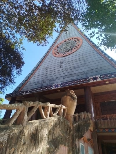This is a ground-level shot of a building in An Phu Ward, Vietnam. The building features a triangular facade made of gray wood planks, decorated with geometric patterns along the edges. A circular emblem is set into the triangle near its apex. Below the facade is a wooden eave decorated with star-shaped patterns. The facade is supported by dark brown wooden columns.
In the foreground, there is a textured cement railing with a large vase-shaped cement post. The railing appears to lead up to the building's entrance. Some tree branches with leaves are visible in the top portion of the frame, framing the building and sky. The sky is a clear blue. The shot is taken in daylight.