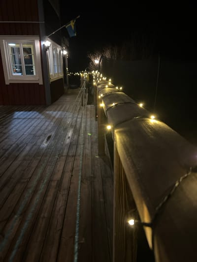 This nighttime image depicts a section of a wooden deck or walkway adjacent to a red-painted building in Gränsta, Sweden. The wooden building, on the left, features white-framed windows with interior illumination and two exterior wall-mounted sconce lights. A Swedish flag is mounted on the building's exterior between the lights. The wooden walkway extends towards the background, illuminated by string lights wrapped around a low wooden railing on the right side. Another light fixture, possibly a lamp post, is visible further along the walkway on the right. A rope or thin chain-like object runs along the left edge of the walkway. The foreground features weathered wooden planks. The background is dark, with silhouetted vegetation visible against the night sky beyond the railing and walkway. No individuals are present.