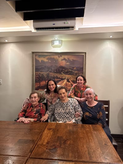 The image shows five women posing for a photo at a wooden table indoors, likely in a dining room. Above them, there is a painting and an air conditioning unit mounted near the ceiling. The women are arranged with three sitting at the table and two standing behind them. The setting is suspected to be in Tijuana, Mexico, but there are no explicit elements in the image to confirm the location.
