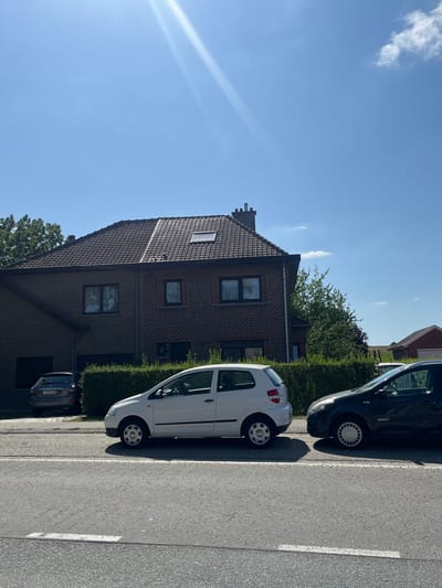 This image depicts a residential street scene in Sint-Pieters-Leeuw, Belgium, under a clear, sunny sky.
In the immediate foreground, a grey asphalt road is visible, marked with two parallel white lines on the left side, indicating a lane boundary or parking area.
The midground features three parked vehicles. Closest to the viewer, positioned on the right side of the frame, is the front section of a dark-colored (likely black) four-door hatchback, possibly a Renault Clio, facing left. Its front passenger-side wheel with a light-colored hubcap is visible. Directly behind this vehicle, and slightly to the left, is a white two-door hatchback, resembling a Volkswagen Fox, facing right. All four wheels are visible, each with a silver hubcap. Both vehicles appear to be parked parallel to the road. Further back and to the left, within a paved driveway adjacent to a house, a dark grey five-door hatchback or small sedan, consistent with a Honda Civic, is parked facing away from the viewer.
The background is dominated by a two-story brick house with a dark brown tiled gabled roof. The roof features a single chimney stack on the right side and a rectangular skylight on the front slope. The house has multiple windows with dark frames. A well-maintained green hedge runs along the front of the property, partially obscuring the lower portion of the house and separating it from the road. To the far right, beyond the house, a small outbuilding with a reddish-brown roof and what appears to be an open field or agricultural land are visible under the expansive blue sky. The sky itself is clear with minimal, wispy clouds. A prominent sun glare emanates from the upper left quadrant of the image. No individuals are visible within the frame.