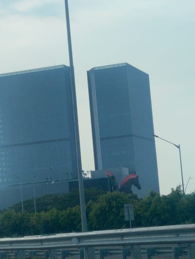 The image captures a view of two modern skyscrapers with a light blue, glass facade, possibly located in Kosambi Barat, Indonesia. The sky is overcast and bright. In the foreground, a metal guardrail runs horizontally across the bottom of the frame. Behind the guardrail, a dense line of green trees is visible. Between the trees and the skyscrapers, a portion of a large, dark structure with a bright red, horse-shaped sculpture is partially obscured. The sculpture appears to be a horse's head and neck with a red mane. Two lampposts are visible in front of the buildings, with one closer to the right side of the frame. A tall, slender pole, possibly a flagpole or antenna, stands vertically between the two skyscrapers and slightly to the left of the horse sculpture. The overall impression is an urban landscape with a distinctive artistic element.