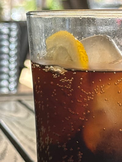 A close-up, eye-level shot shows a glass of dark brown soda with ice and a lemon slice. The glass is transparent and has some condensation on the exterior. The soda is fizzy, with numerous small bubbles rising to the surface. The lemon slice is a bright yellow wedge, perched on the rim of the glass. The background is blurred, with hints of green foliage and what appears to be a dark, textured surface, possibly a table. The setting appears to be outdoors, on a sunny day, in the city of Campillo del Moro, Spain.