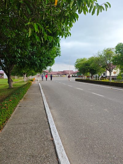 The image depicts a paved road with a sidewalk on the left, bordered by lush green grass and trees. Several individuals are walking on the sidewalk and on the road. Buildings with red roofs and white walls are visible in the background. The sky is overcast. The scene appears to be a tranquil, well-maintained area, possibly a park or a residential complex within the city of Glokaron, Indonesia.