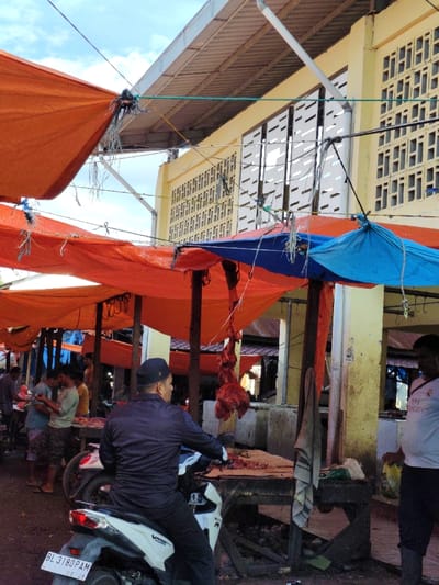 A bustling outdoor meat market with rows of orange and blue tarp-covered stalls. A man on a white scooter, bearing license plate BL 3180 PAM, is stationary near a butcher's stall displaying hanging raw meat. Several people are present, either standing near the stalls or engaging in conversation. The market appears to be adjacent to a two-story building with perforated window coverings.