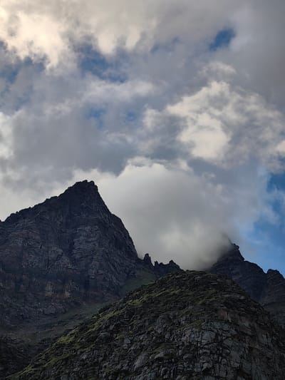 The image shows a dramatic mountain landscape under a cloudy sky. In the foreground, a rocky, green-covered slope rises steeply. Behind it, jagged, dark grey mountain peaks pierce through white and grey clouds. Some of the peaks are sharp and pointed, while others are partially obscured by fog. Patches of blue sky are visible between the clouds. The lighting suggests it is either early morning or late afternoon. No specific landmarks indicating the city of Damphug, India are visible in this image.