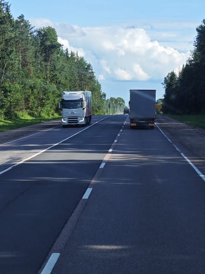 An eye-level, wide shot captures a clear, sunny day with a bright blue sky dotted with puffy white clouds. The scene depicts a two-lane asphalt road winding through a dense forest. On the left side of the road, a white semi-truck travels in the left lane. On the right side of the road, a dark-colored semi-truck is ahead of the white truck and is traveling in the right lane. The trees on both sides of the road are lush and green, indicating a summer season. White lane markings are visible on the road, dividing the lanes and marking the shoulders. Shadows from the trees stretch across the asphalt, dappling the surface with light and dark. The overall impression is one of peaceful, rural travel.