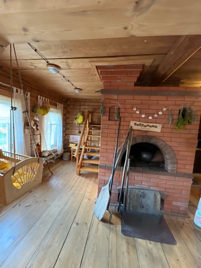 In a rustic, wooden interior in Vybolovo, Russia, a traditional Russian stove, or "pech," dominates the scene. Constructed of red brick, the stove features an arched opening containing a dark, cast-iron pot. Above the opening, a wooden sign reads "Бабушкино Подворье" (Grandmother's Courtyard), adorned with strings of dried herbs and garlic. To the left of the stove, a wooden scoop and a shovel rest on a metal ash pan. A wooden staircase leads upwards, partially obscured by hanging decorations. To the left of the stairs, a window with white curtains overlooks an outdoor scene. Near the window, a wooden cradle with yellow bedding is suspended by ropes. The room is furnished with wooden floors and log walls, and several decorative items, including stuffed toys and dried bunches of herbs, are visible. Lighting fixtures are mounted on the wooden ceiling.