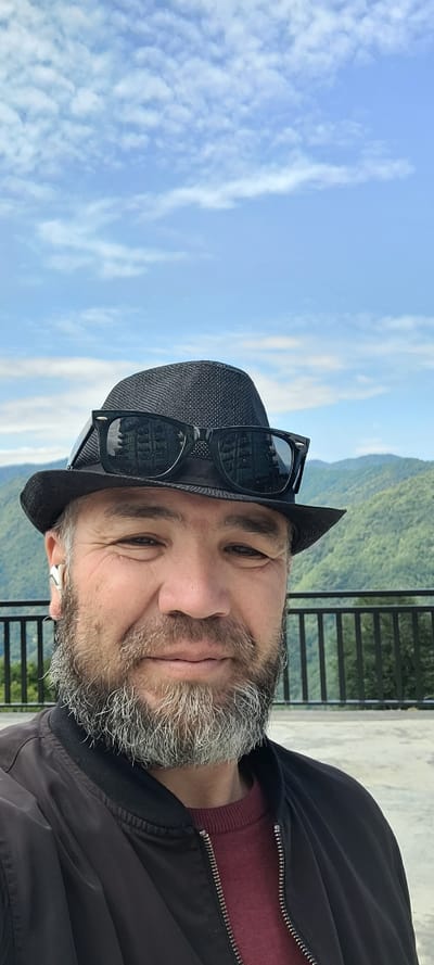 A man in a black fedora hat and sunglasses takes a selfie outdoors. He has a gray beard and is wearing a black jacket over a maroon sweater. Behind him, a black metal railing separates him from a lush, green mountain landscape under a bright blue sky with scattered white clouds. The location is Milisi, Georgia.