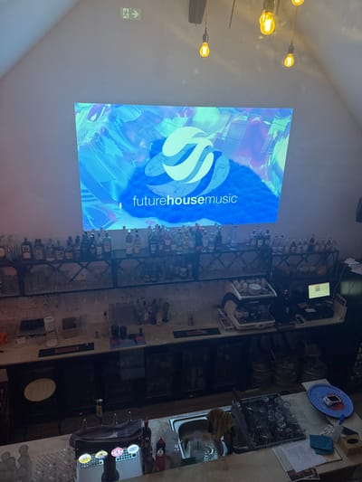 This image, captured inside a venue in Dambach-la-Ville, France, presents an elevated view of an unoccupied bar area, characterized by a sloped ceiling and white walls. A large projection dominates the back wall, displaying a blue-toned graphic with a white, stylized, multi-curved logo and the text "futurehousemusic." Overhead, industrial-style pendant lights with exposed bulbs illuminate the space, and a green exit sign is visible on the upper left wall.
Below the projection, a two-tiered metal shelving unit holds numerous bottles of spirits and liquors. Directly beneath this, the main bar counter features a white and chrome espresso machine, a point-of-sale system with a monitor displaying green text, and various bottles and glassware. The lower section of the counter reveals beer taps, a stainless steel sink with a faucet and a yellow cloth, a large plastic tray filled with inverted clean glasses, and a blue plastic tray holding a small electronic device. Below the main counter, dark refrigeration units are visible. No individuals are present in the scene.