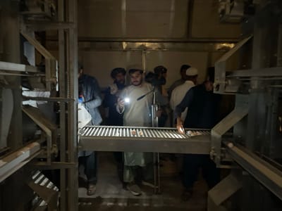 The image depicts an interior view of an egg production or processing facility in Shuyene Vosta, Afghanistan, characterized by multi-tiered metallic machinery with conveyor belts. Approximately seven adult males are present, gathered around a horizontal conveyor belt displaying multiple light-colored eggs. The environment is dimly lit, requiring the use of handheld artificial light sources. A central male, wearing a light-colored tunic, illuminates the eggs on the conveyor with a white flashlight held in his right hand. Another male to his right, wearing a dark vest over a lighter garment, also holds a light source and is reaching towards the eggs on the conveyor. Other males, dressed in varied traditional and casual attire, observe the scene from various positions around the machinery. The actions indicate inspection, collection, or maintenance of the egg processing line under low-light conditions.
