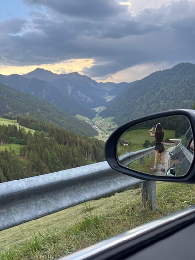 This media file is a vertical, eye-level shot of a mountain landscape as seen from the perspective of a car. The primary focus is a side-view mirror reflecting a young woman with long brown hair, wearing a brown outfit and holding a phone, seemingly taking a picture. She is standing on the grassy shoulder of a road, with a guardrail to her right.

The reflected scene in the mirror reveals a verdant valley with scattered houses and green fields, bordered by dense pine forests on the slopes of surrounding mountains. The sky is partly cloudy, with dramatic streaks of sunlight breaking through the clouds, suggesting it might be late afternoon or early evening. The overall mood is one of scenic travel and personal exploration. The location is identified as Ratschings - Racines, Italy. There is no visible text in the image.