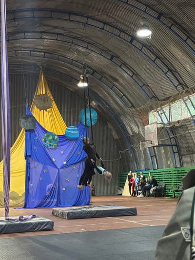 A group of people, likely performers, are suspended in the air within an indoor gymnasium. Two individuals, one dressed in a dark outfit and another in a teal and black costume, are the most prominent subjects. They appear to be performing an aerial act, possibly on silks or a trapeze, indicated by the fabric and rigging visible. They are positioned above a large, draped fabric structure that resembles a celestial scene with blue fabric adorned with stars and yellow fabric. Several spherical objects, resembling planets, are suspended around the performers, further enhancing the cosmic theme.

The setting is a large, industrial-style gymnasium with a corrugated metal ceiling and exposed beams. Lights are suspended from the ceiling, illuminating the performance area. In the background, a basketball hoop is visible, suggesting the space is also used for sports. Spectators are seated on green bleachers to the right, observing the performance.

The image appears to be taken indoors during an event, possibly an evening performance, given the artificial lighting. There are no immediate clues about the weather or time of day outside the venue. The overall mood seems to be one of active performance and observation. No visible text is present in the image. The context provided places the location in Pribrezhnyi, Russia.