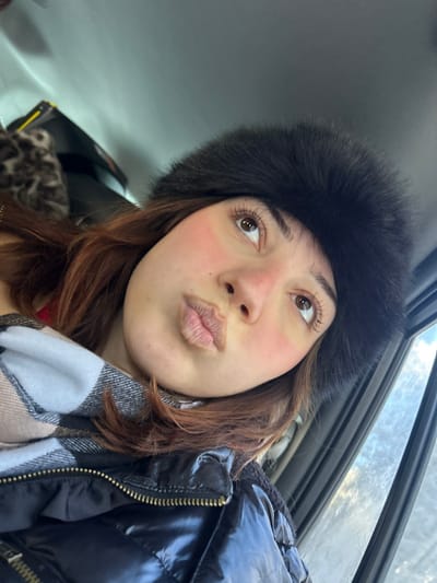 An individual is depicted in a close-up, high-angle selfie photograph taken inside what appears to be a vehicle, in or near Le Hohwald, France. The subject is a female with reddish-brown hair, dark brown eyes, and prominent eyelashes. She is wearing a dark, fluffy winter hat, a light-colored plaid scarf, and a black, shiny quilted puffer jacket with a visible zipper. Her cheeks exhibit a rosy flush, and her lips are pursed into an expressive shape. The immediate background shows a light grey ceiling to the left, with a partial view of a dark object featuring a leopard print texture. To the right, a window frame is visible, with a small portion of a bright, possibly overcast, sky discernible through the window.
