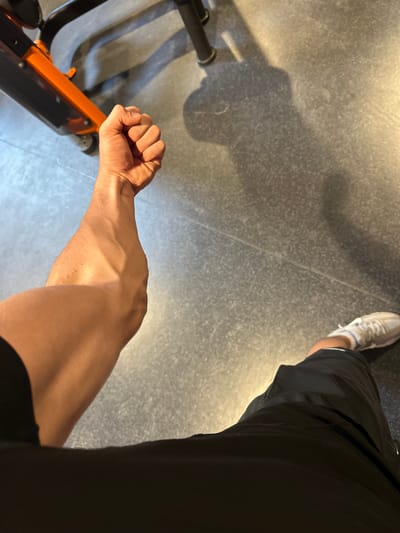 A person's arm is clenched into a fist and extended forward. The person is wearing black shorts and white sneakers. The scene appears to be in a gym, with the shadow of the person's arm cast on a textured gray floor. An orange and black exercise machine is partially visible in the background. There is no indication of the location in Alès, France.