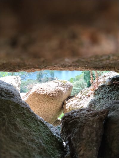 The image shows a view through a narrow gap between large rocks. The foreground is dominated by the rough, textured surfaces of the stones, some covered in moss. Beyond the gap, there is a glimpse of greenery, trees and a body of water in the background. The scene is set outdoors. The location is in Alijó, Portugal. There are no people or animals present in the scene.