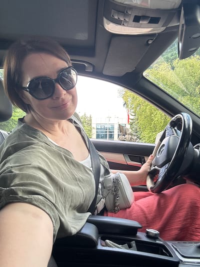 A woman with short brown hair and sunglasses is sitting in the driver's seat of a car, holding the steering wheel. She is wearing a green top and a pink skirt. A white purse is hanging from her shoulder. In the background, through the car window, a building with a red flag and statues can be seen. The car's interior features a black dashboard, a sunroof, and a center console with a gear shift knob. The setting appears to be in a city, possibly Etimesgut, Turkey, given the presence of the building and statues. The image is a selfie taken from inside the car.