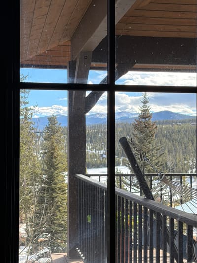 A view through a window reveals a mountain landscape with snow and trees. An architectural structure featuring beams and siding is on the left. A deck with a metal grate and some kind of metal chair are visible on the right. The image is horizontally oriented.