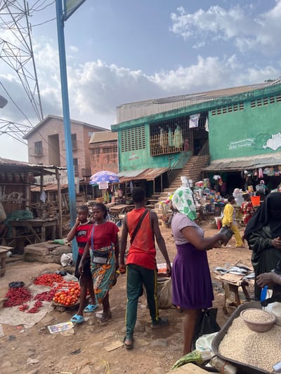 The image shows a street market in Ondo, Nigeria, located at latitude 7.253 and longitude 5.195. There are six people present, including three women and three men. The women are all wearing traditional clothing. Two of the women are wearing headscarves, one in a white floral pattern and one in black. The men are all wearing t-shirts and trousers. One of the men is wearing a red t-shirt, another is wearing a black t-shirt, and another is wearing a white vest. A table displays red peppers and tomatoes in the foreground. Other tables in the background display fish, grains, and other goods. There are several buildings in the background, including a building with a sign that reads "Cholesterol FREE ZERO TransFAT". The ground is dirt and there are several power lines overhead. The sky is cloudy. There are multiple stalls on either side of the street, with people moving between them. A woman in a purple dress is walking between stalls and looking to the side. A man in a red shirt is walking in the opposite direction. A woman wearing a black headscarf is holding a tray of white grain.