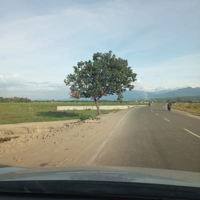 The image shows a view from inside a vehicle, looking out onto a road in Dayah Kruet, Indonesia. A tree stands to the left of the road, with a field and low wall visible in the background. Two motorcycles are on the road in the distance, heading in the same direction as the vehicle. Mountains are faintly visible in the far background under a partly cloudy sky. The shoulder of the road on the left side appears unpaved and uneven.