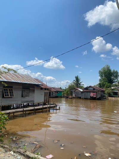 In the city of Berangas Timur, Indonesia, a body of brown, murky water reflects a bright blue sky dotted with fluffy white clouds. Along the water's edge, a series of weathered wooden stilt houses, some with rusty tin roofs, are clustered together. A rickety wooden walkway connects some of the houses, providing passage over the water. The water is littered with various types of debris, including plastic bags, bottles, and other refuse, indicating a degree of pollution. Green foliage frames the scene on the left, while trees are visible in the background. The overall impression is of a humble, waterside community with signs of environmental neglect.