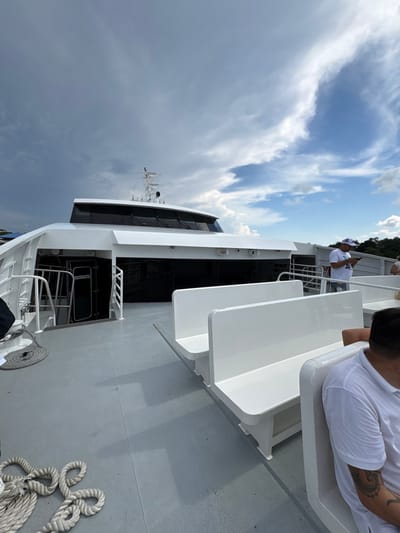 The image displays the open upper deck of a white passenger catamaran ferry, located in the city of La Ceiba, Honduras, likely at its port. The deck surface is grey. In the foreground-left, a thick white mooring rope is coiled on the deck. A white staircase with handrails descends into the vessel's interior through an open dark doorway on the port side.
Rows of white, molded plastic bench seats are visible on the deck; two rows are unoccupied in the mid-ground, while the back of an occupied row is in the immediate foreground on the right. A male individual is seated in this foreground row, with his right arm, featuring a tattoo, visible wearing a white short-sleeved shirt. Further along the starboard side, a male figure stands wearing a white baseball cap, white t-shirt, and jeans, looking down at a mobile phone. A third individual is partially visible further back on the starboard side.
The vessel's forward superstructure is white with dark, tinted windows for the bridge area. A central mast equipped with antennae and navigation equipment rises above the bridge. The sky overhead is partly cloudy, with dark grey storm clouds dominating the left side, transitioning to lighter white clouds against a blue sky on the right. A distant landmass is visible on the far-right horizon.