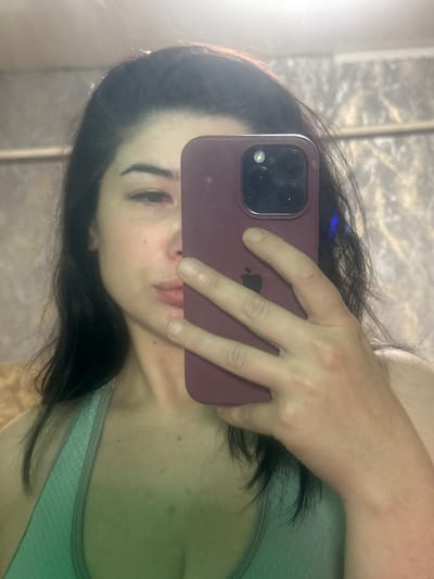 A person is taking a selfie in a mirror. The person has long, dark hair and is wearing a light-colored sports bra. They are holding a maroon smartphone with a camera array in their right hand, which is obscuring their face from the nose up. The background appears to be a bathroom with a patterned wall and a shelf. There is a green hue cast over the lower part of the image. The image does not provide any specific visual cues to determine its location in the city of Gavrilov Posad, Russia.