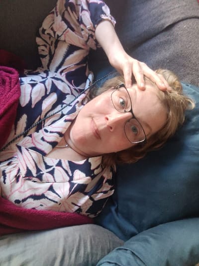An indoor scene in Libramont, Belgium, depicts a light-skinned individual, appearing female, lying on a dark gray sofa or couch. The individual's head rests on a dark blue pillow, positioned towards the upper right of the frame. She wears dark-framed glasses and a collared shirt with a pattern of dark blue and pinkish-white abstract leaf shapes. A silver-colored chain necklace is visible around her neck. Her right hand is raised, with fingers resting on her forehead near the hairline. A dark pink or magenta blanket partially covers her left side and torso, while a light gray fabric, possibly trousers or another blanket, covers her lower body. The individual's eyes are open and directed towards the camera, with a neutral expression. The background is out of focus, indicating an interior setting.