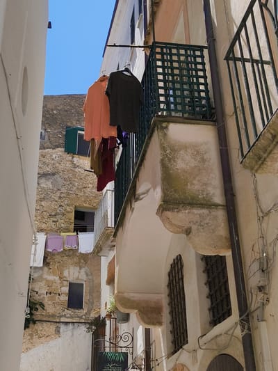 The image captures a narrow alleyway in Sperlonga, Italy, under a clear blue sky. Laundry hangs on clotheslines strung between buildings. On the left, a white building dominates the foreground, with a curved wall and a circular indentation. To the right, a peach-colored building has a balcony with a dark green lattice railing. Several articles of clothing, including what appear to be two t-shirts (one peach, one dark gray), are hanging on a clothesline in front of this building. Further to the right, a building with a textured, light-colored facade features a dark iron railing and a downspout. Another balcony, also with an iron railing, is visible above. In the background, a stone building with a green-shuttered window is visible. Below, a gate with ornate scrollwork and a sign that reads "THE ROOTS" is present. The architecture suggests a traditional Mediterranean style, common in coastal Italian towns. There are no people visible in the scene.