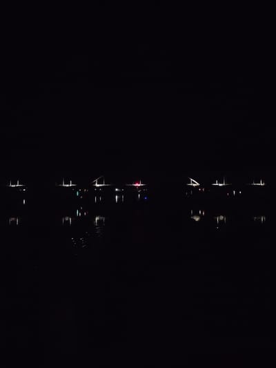 The image shows a nighttime scene with several boats on a body of water. The lights of the boats reflect on the water's surface. The boats appear to be relatively small, possibly sailboats or yachts. The scene lacks specific details, making it difficult to determine a specific location, like Zarya, Russia, or specific activities. The ambient lighting is very low, with the primary light sources being the boats' lights and their reflections.