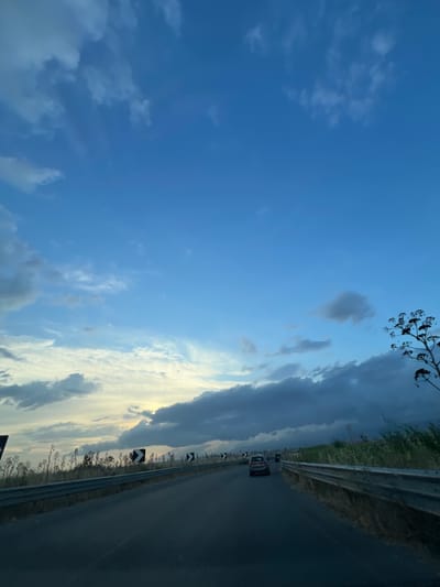 The image captures a view from a vehicle traveling on a road in Manfredonia, Italy, under a dynamic sky. The road curves gently to the right, bordered by a metal guardrail on both sides. The left side of the road is lined with dry, tall grasses and thin, bare trees, with several chevron warning signs indicating curves. The right side of the road also features vegetation and a few of the same chevron signs.

The sky is a dominant feature, transitioning from a bright, pale blue near the horizon with patches of white and gray clouds, to a deeper blue with scattered, darker clouds higher up. The sun appears to be setting or has recently set, casting a warm glow in the distance behind the clouds, creating a dramatic contrast.

Two cars are visible on the road ahead, one in the left lane and another further ahead in the right lane. The atmosphere suggests it is late afternoon or early evening. The scene is devoid of people other than those within the vehicles. The environment appears to be semi-rural, possibly on the outskirts of the city, with the road leading towards a distant, hazy landscape.