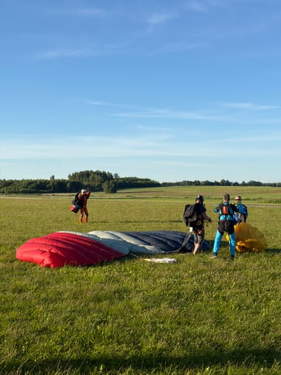 In a wide, grassy field under a clear blue sky with scattered wisps of clouds, a group of skydivers are preparing for or have just completed a jump. In the foreground, a large, folded parachute lies on the grass, its fabric a vibrant mix of red, white, and blue. To the right of the parachute, a bright yellow bag, likely containing another parachute, rests on the ground.

Several skydivers are visible in the mid-ground. One skydiver, dressed in an orange and black jumpsuit, is walking away from the camera towards the left of the frame, holding a parachute pack. Further to the right, three skydivers are interacting near the yellow bag. Two are wearing blue jumpsuits, and one is wearing a black top and grey pants with a black backpack. The skydiver in the black top appears to be adjusting rigging on a parachute.

The background features a line of lush green trees bordering the field, with more trees and distant landscape visible under the expansive sky. The scene suggests an outdoor airfield or drop zone, typical for skydiving operations. Based on the vegetation and general landscape, this location is consistent with an open field area found in or around the city of Limbaži, Latvia. The lighting indicates it is daytime, likely late afternoon given the warm tones and long shadows.