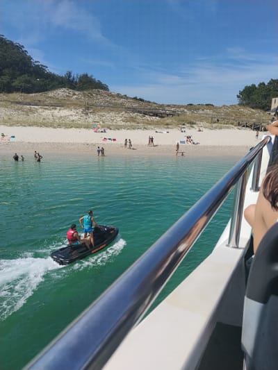 In Vigo, Spain, on a sunny day, two individuals are riding a jet ski on the turquoise waters of the Atlantic Ocean. The jet ski, a black Sea-Doo GTX, is moving away from the viewer, creating a white wake. The driver wears a blue life vest and blue shorts, while the passenger in front wears a red life vest and white shorts.
In the background, a sandy beach stretches along the coastline, backed by dunes covered in sparse vegetation. Wooden walkways are visible on the dunes, leading towards the beach. Several people are scattered across the beach, some sunbathing, others standing or walking. A few individuals are in the shallow water near the shore.
The foreground of the image is dominated by the railing and side of a white boat, suggesting the photo was taken from a larger vessel. The sky above is a clear, bright blue with a few wispy white clouds. The overall scene depicts a vibrant summer day at the beach with recreational activities taking place in the water and on the shore.