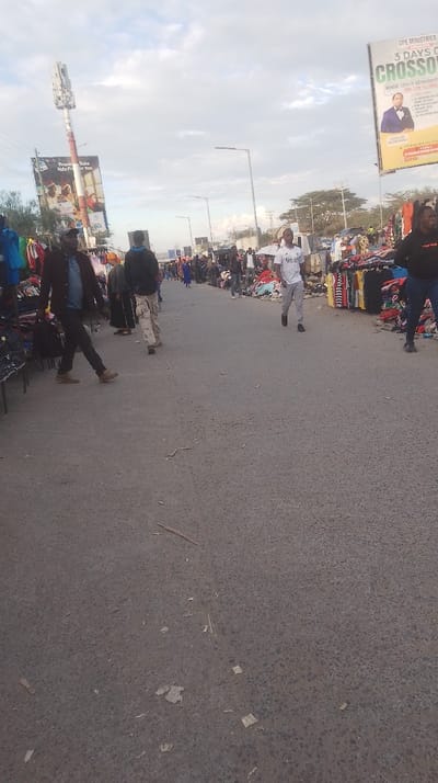 The image captures a bustling outdoor market scene along an asphalt road in Oloosirkon/Sholinke ward, Kenya. The setting appears to be a street or open area where vendors have set up stalls on both sides, displaying various merchandise, predominantly clothing, on racks and tables. Numerous people are present, walking along the road, some appearing to browse or shop, while others might be vendors or passersby.
Subjects include men and women of various ages. Several individuals are clearly visible, including a man in a dark jacket and trousers walking on the left foreground, another man in a white t-shirt with a "Real Madrid" logo walking in the mid-ground, and a woman in dark attire on the right. The road itself is gray asphalt, with some debris scattered on its surface. In the background, more market activity can be seen, with additional people and stalls extending into the distance.
Notable details include two large billboards. The one on the left features text "Present Yako Ndio Pesa Yako" (Your Presence Is Your Money) and "Safiri," likely related to Safaricom, with images of people. The billboard on the right advertises "CPE MINISTRIES PRESENTS 3 DAYS OF CROSSOVER" with dates "MONDAY 29TH TO WEDNESDAY 31ST" and displays a picture of a man in a suit. A tall communication tower is visible behind the left billboard. Streetlights line the road. The sky is overcast with light clouds, suggesting either early morning or late afternoon light, not direct harsh midday sun. The overall atmosphere is active and typical of a local market.