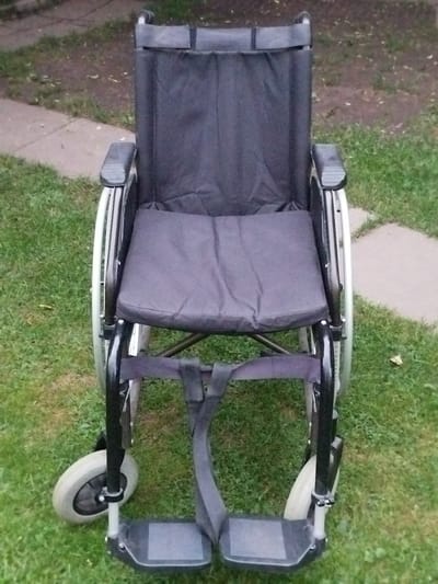 An empty, dark gray fabric wheelchair is positioned outdoors on a patch of green grass. The wheelchair has black armrests, a black seat cushion, and black footrests. Its large rear wheels are partially visible on either side, and smaller front wheels are present. A path made of light-colored rectangular stones is visible to the left of the wheelchair, and the background consists of more grass and some fallen leaves. The scene appears to be in a park or garden setting.
