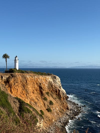 The image depicts a scenic view of Point Vicente Lighthouse in Rancho Palos Verdes, United States of America. The lighthouse, a white tower with a black top, stands on a cliff overlooking the Pacific Ocean. A palm tree stands nearby. The coastline features a rocky cliff with shrubs and vegetation. The ocean stretches to the horizon under a clear, blue sky. The waves crash against the rocks.