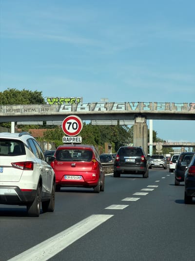The image shows a traffic jam on a highway, likely in Bron, France, given the license plates and signage. Several cars are visible, including a white SUV in the foreground on the left, a red Kia hatchback, and a black SUV. The cars are on a multi-lane highway under a clear blue sky. Above the highway, a bridge or overpass is covered in graffiti. A speed limit sign indicates "70" and the word "RAPPEL" (reminder) below it. The highway has painted lane markings, including a solid white line on the left and dashed white lines separating lanes. The environment appears to be urban or suburban, with trees visible in the background. The overall scene depicts a common traffic situation with a sense of congestion.