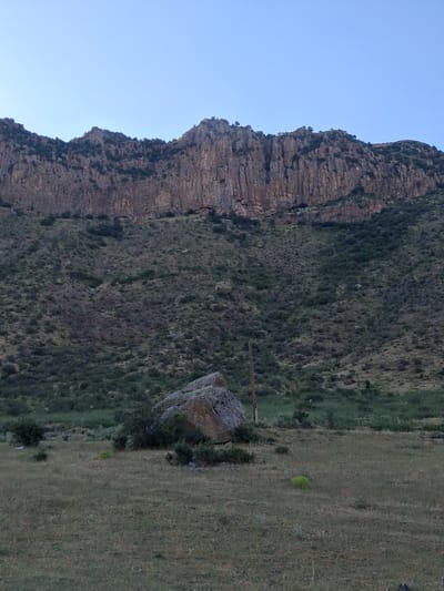 This media file captures a dramatic mountain landscape in Shatin, Armenia. The scene is dominated by a sheer, rocky cliff face that rises steeply, showing distinct layers and textures of the geological formations. The cliff is partially covered in sparse green vegetation, adding a touch of life to the rugged terrain.
To the right of the cliff, a grassy slope extends outwards, with dry grass suggesting a warm, possibly dry season. Scattered across the grassy area are a few small bushes and a prominent, large boulder resting near the transition between the vegetated slope and the grassy field. A vertical wooden pole stands in the middle ground, suggesting it might be part of a fence or some other marker.
The sky is a clear, pale blue, indicating it is daytime, likely early morning or late afternoon given the soft lighting and lack of harsh shadows. There are no visible people or animals in the frame. The overall impression is one of natural grandeur and solitude, highlighting the raw beauty of the Armenian highlands. There is no visible text in the image.