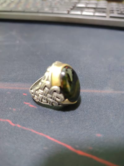 The image is a close-up, slightly blurry shot of a silver ring with a dark, possibly obsidian, stone set in it. The ring has an intricate, woven-like design on the band. The ring is resting on a dark, textured surface, possibly a desk or mousepad, with faint red lines visible. In the background, out of focus, is a keyboard. There are no people or actions depicted in the image, and the environment is indoor. The image does not provide any information related to Sariasiya, Uzbekistan.