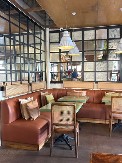 The image depicts the interior of a restaurant, possibly in Mazatlán, Mexico, given the architectural style and furnishings. The seating arrangement features a long, brown leather banquette along the wall, adorned with various throw pillows in neutral tones. Individual wooden chairs with woven backs are placed along the banquette, each paired with a small, light-green-topped table. The chairs are supported by black, ornate cast-iron bases.
Large, gridded windows run along the wall opposite the banquette. The windows appear to be reflecting the interior of the restaurant, as well as elements of the outside environment, including greenery and other buildings. The lighting consists of multiple white, cone-shaped pendant lights hanging from the ceiling.
The overall impression is of a modern, stylish restaurant with an industrial-chic aesthetic. There are reflections visible in the windows, suggesting the presence of people within the restaurant, though they are not clearly discernible. The image suggests a casual yet sophisticated dining environment.