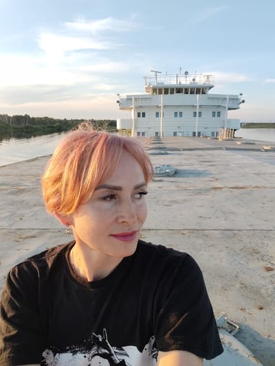 The media file shows a woman with short, pinkish hair, wearing a black t-shirt, and earrings. She is looking to the right, with a slight smile. Her makeup includes mascara and lipstick. The setting appears to be the deck of a large white ship or barge, with a body of water and a tree-lined shore in the background. The sky is partly cloudy with a warm glow, suggesting it is either sunrise or sunset. The large white structure behind her is likely the superstructure of the vessel. The overall mood is peaceful and contemplative. There is some faint, partially obscured graphic text on the woman's t-shirt, but it is not legible. The location is identified as Nifantovo, Russia.