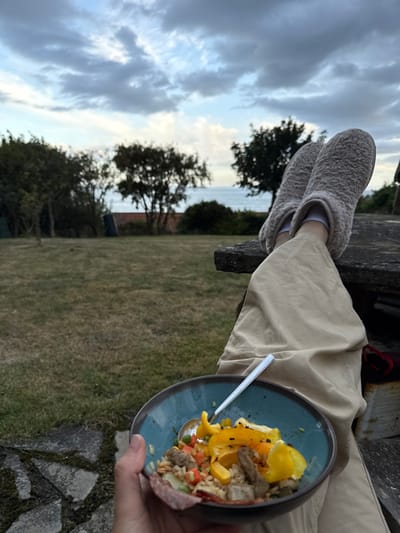 The image displays an outdoor scene in a garden setting in Crail, United Kingdom. In the foreground, a person's left hand holds a teal-colored ceramic bowl with a brown rim, containing a mixed meal composed of rice or grains, diced orange and yellow bell peppers, green peas, diced carrots, meat pieces, and slices of cured sausage positioned on the rim. A silver spoon rests within the bowl. The person's lower legs and feet, clad in light-beige fluffy slippers and light-colored trousers, are elevated and rest upon a dark stone surface to the right of the frame. The mid-ground consists of a green lawn bordered by various shrubs and trees. A red brick wall and a dark container are visible through the foliage. In the distant background, the sea is visible under an overcast sky with scattered clouds, suggesting late afternoon or early evening.