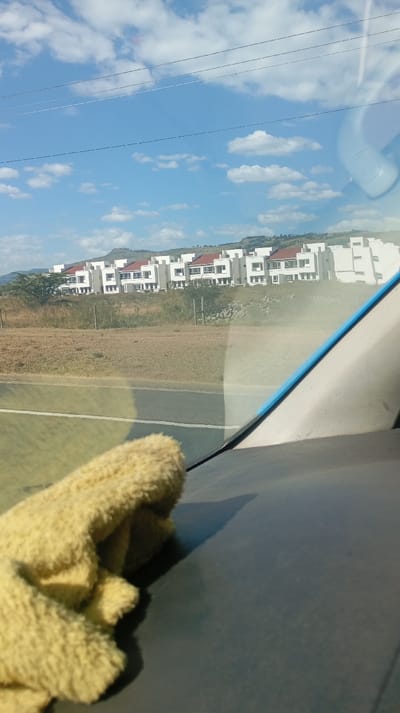 A row of white buildings with red roofs lines the horizon under a blue sky dotted with clouds. They sit atop a slight rise. A road stretches in front of the buildings, with a grassy field between the road and the buildings. The scene is viewed through a car windshield, with a yellow cloth resting on the dashboard. The car's interior is visible, including a portion of the dashboard and the blue trim of the windshield.