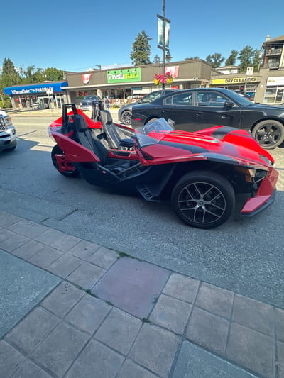 In Port Coquitlam, Canada, a red three-wheeled vehicle is parked on a street in front of a strip mall under a clear blue sky. The vehicle, with its sleek, sporty design, is positioned in the foreground, angled slightly towards the viewer. Behind it, the strip mall houses several businesses including a "PharmaChoice Pharmacy" with a blue awning and a "Freshmart" grocery store with a prominent green sign. A black sedan is parked in front of the dry cleaners. The street is paved, and a sidewalk with square concrete tiles lines the edge of the scene. Trees and buildings are visible in the background, suggesting a typical urban or suburban setting. The overall impression is a sunny day in a commercial area.